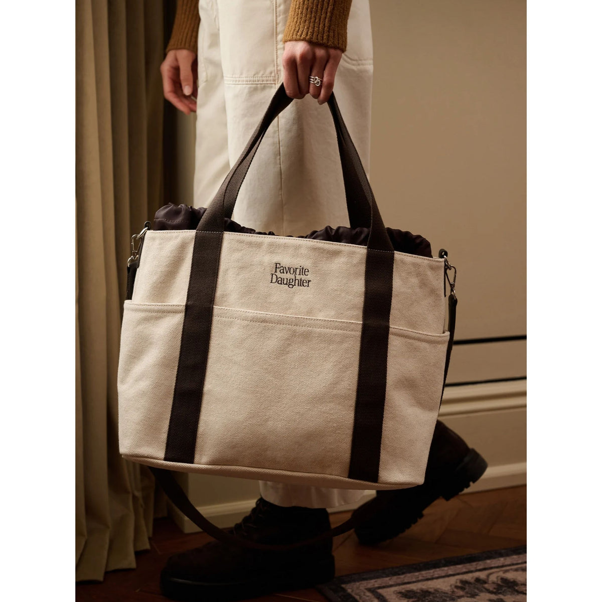 A person holds a large canvas tote bag with dark brown straps and trim. The bag is cream-colored with the text 'Favorite Daughter' printed on the front.
