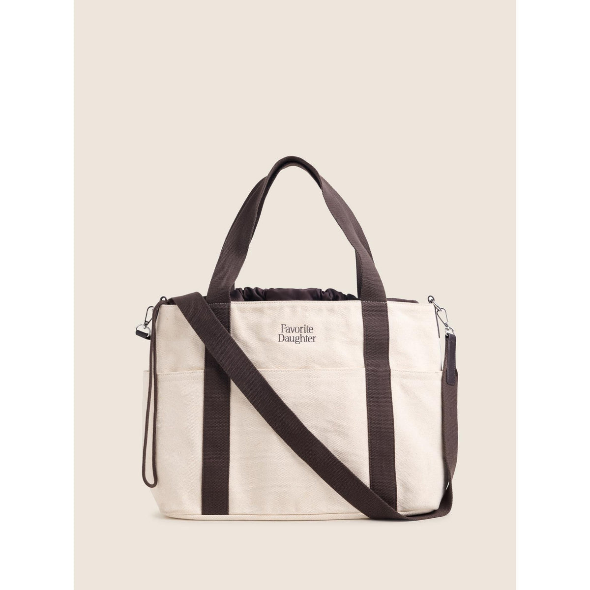 An off-white canvas tote bag with dark brown handles, side straps, and a matching adjustable shoulder strap. The brand name 'Favorite Daughter' is printed in black on the front.