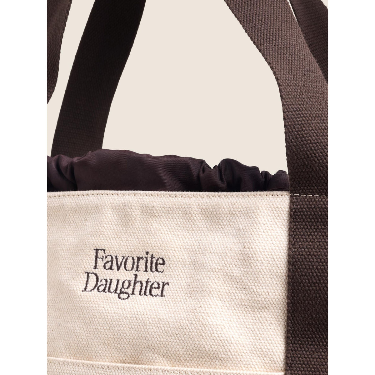 Close-up of a canvas tote bag with dark brown webbed handles and a dark brown gathered top trim. The natural beige canvas body features the text 'Favorite Daughter' printed in dark brown on the front.