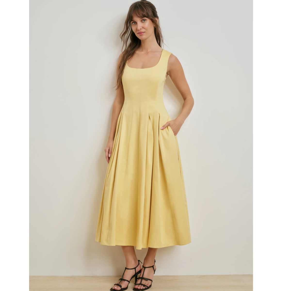 A woman modeling a sleeveless midi dress in a soft pastel yellow, standing against a plain light gray wall.