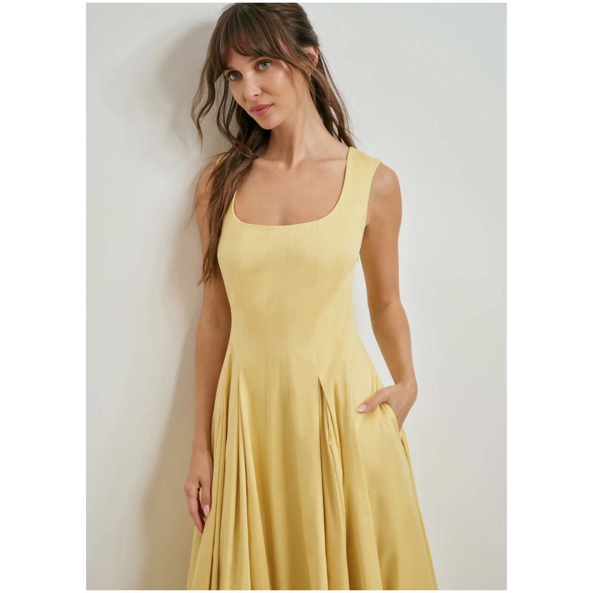 A woman wearing a sleeveless yellow dress with a square neckline and a flared skirt, standing against a plain light gray wall.