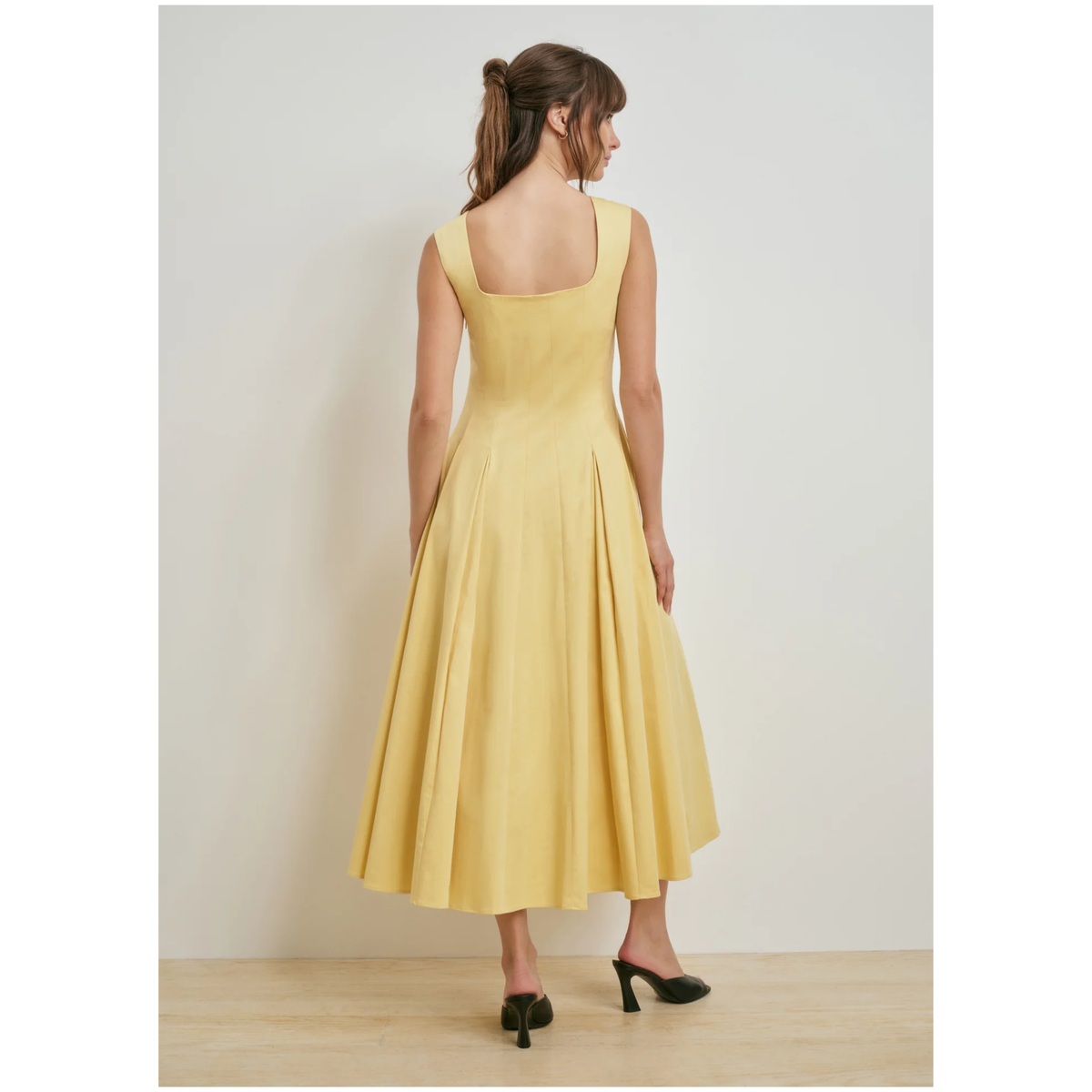 A woman wearing a sleeveless yellow midi dress with a structured bodice and flared skirt, shown from the back against a light gray studio wall.