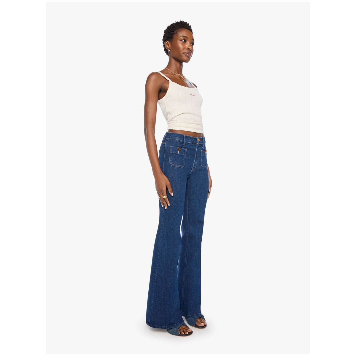 A model wearing a white ribbed tank top and dark blue high-waisted flare jeans, standing against a light gray studio background.