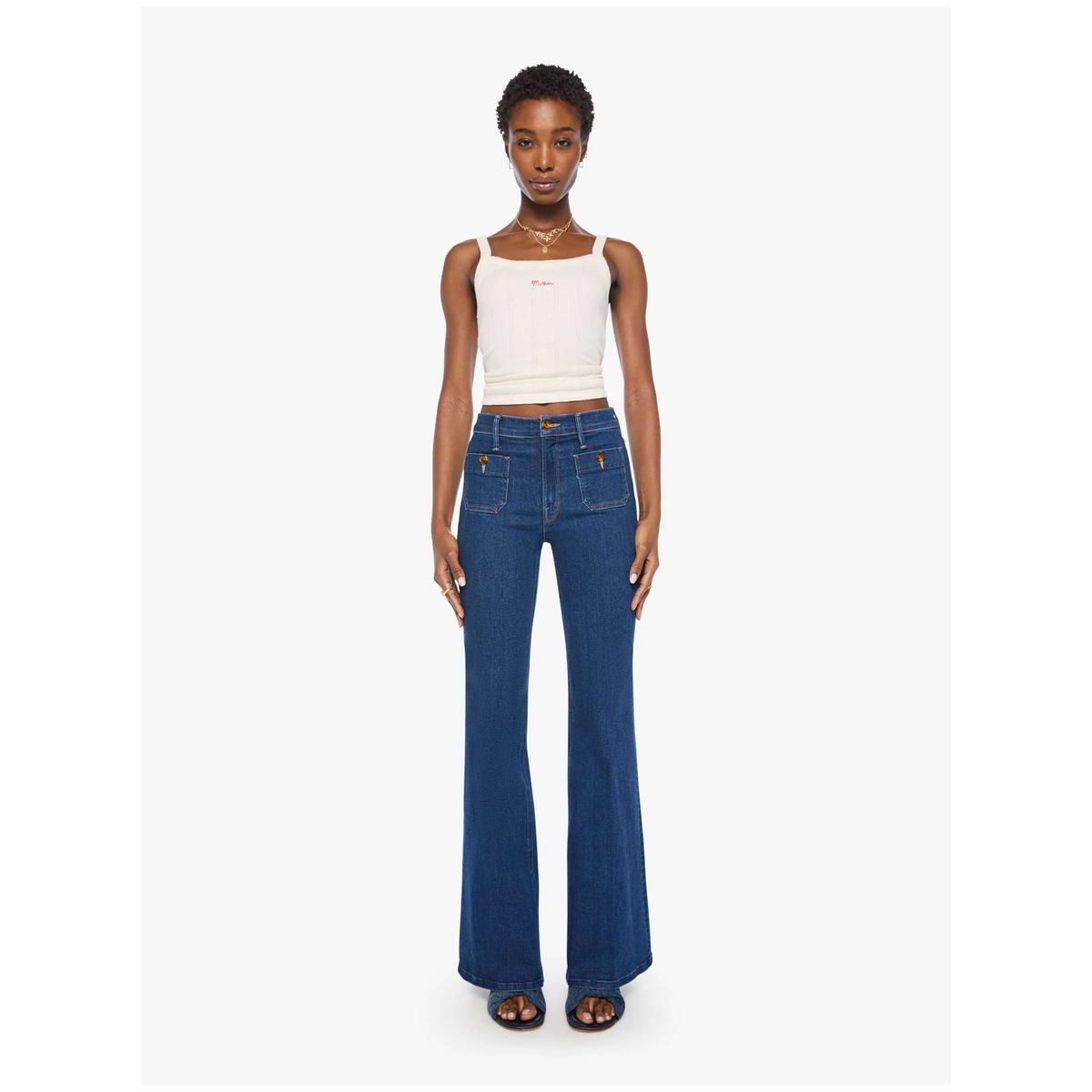 A model stands against a light gray studio background wearing a white cropped camisole top and dark blue wide-leg denim jeans with gold-tone hardware.
