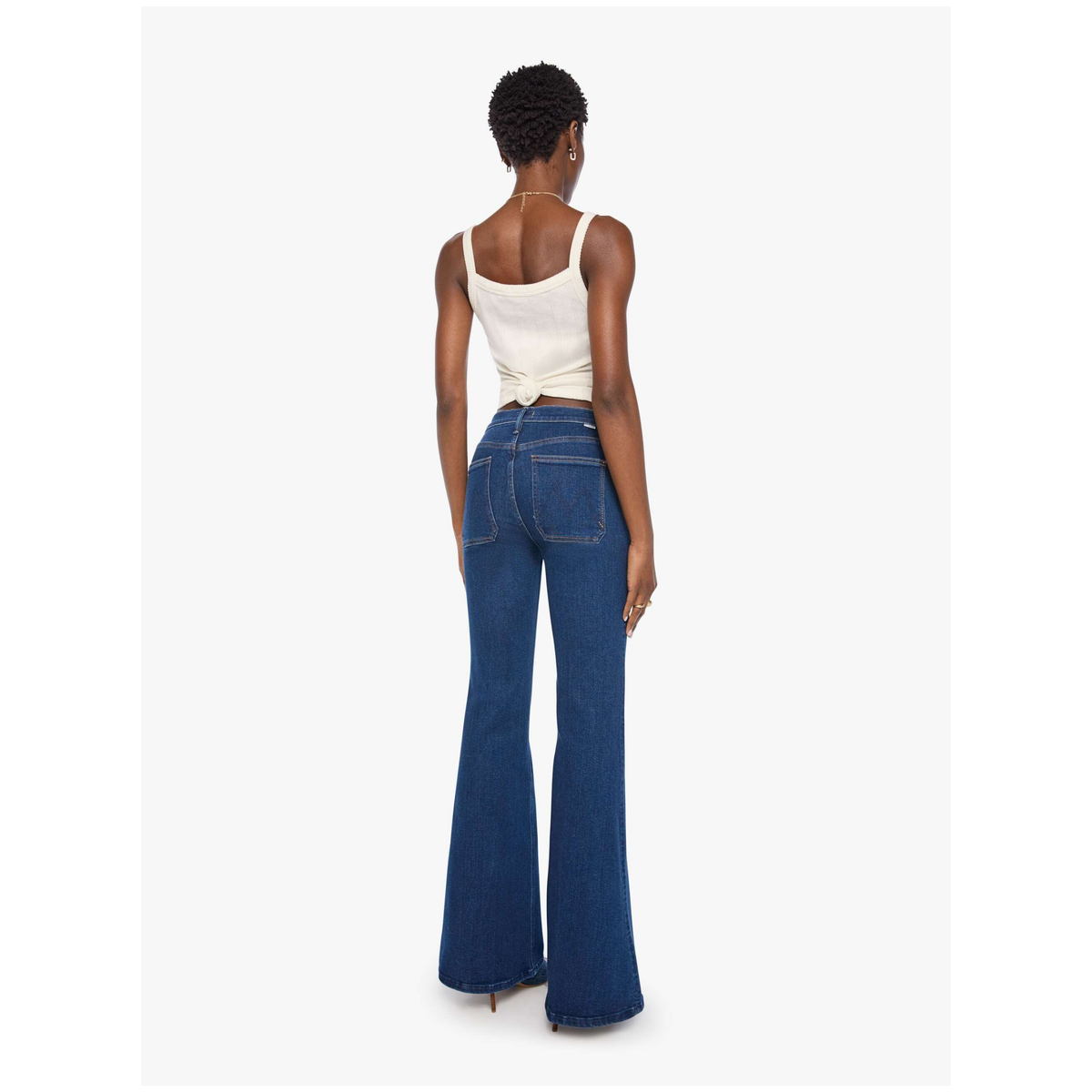 A woman shown from the back wearing dark blue flare jeans and a cream-colored wrap top, standing against a light gray studio background.
