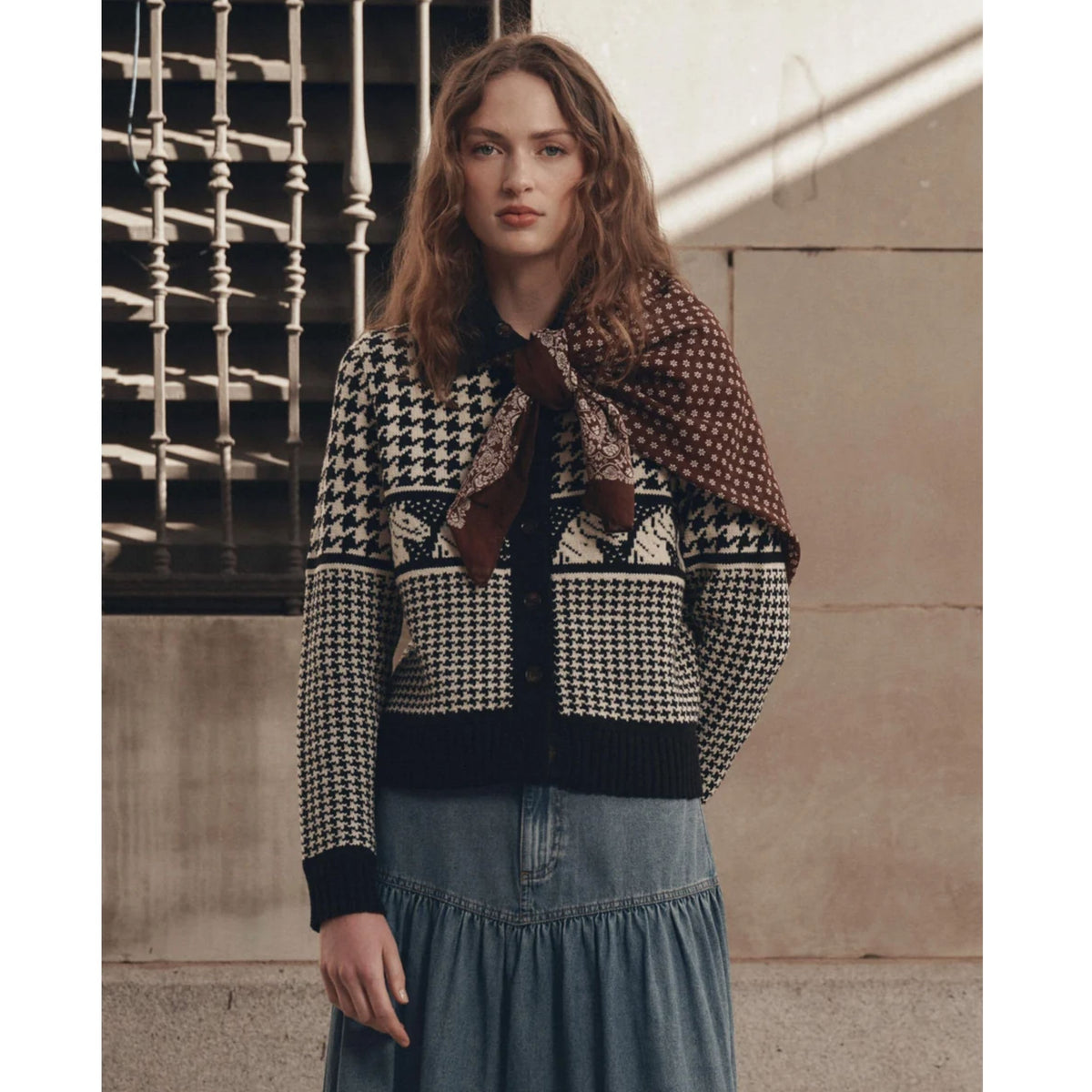 A woman wearing a black and cream houndstooth knit cardigan with a brown patterned scarf tied around her neck, paired with a blue denim skirt, standing against a beige stone wall with a metal railing.
