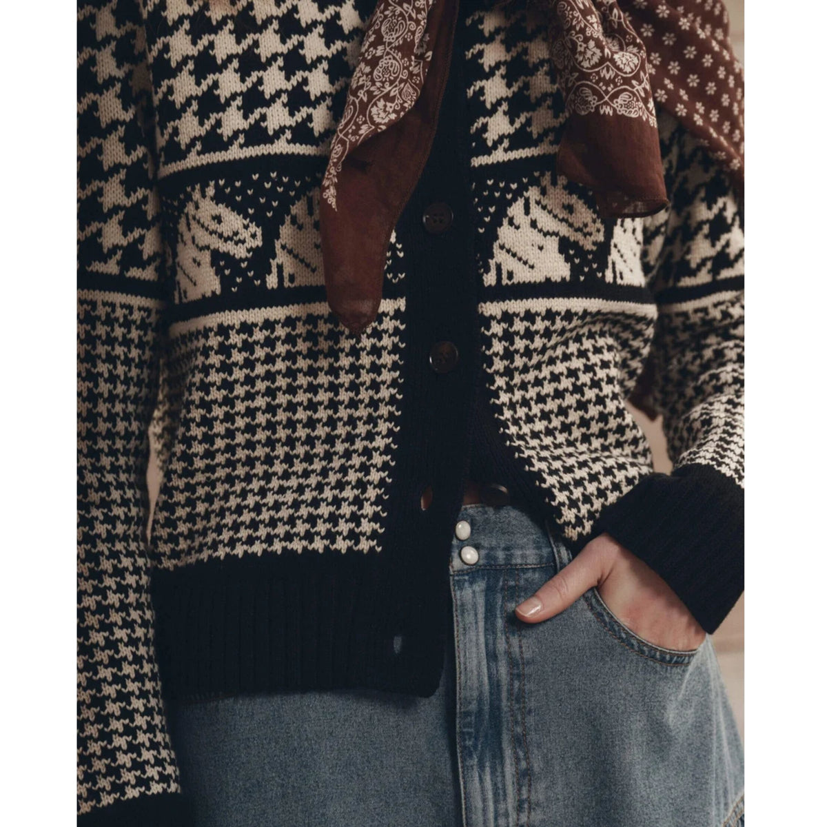 A close-up of a person wearing a black and cream houndstooth patterned cardigan with a brown patterned scarf, paired with light blue denim jeans.