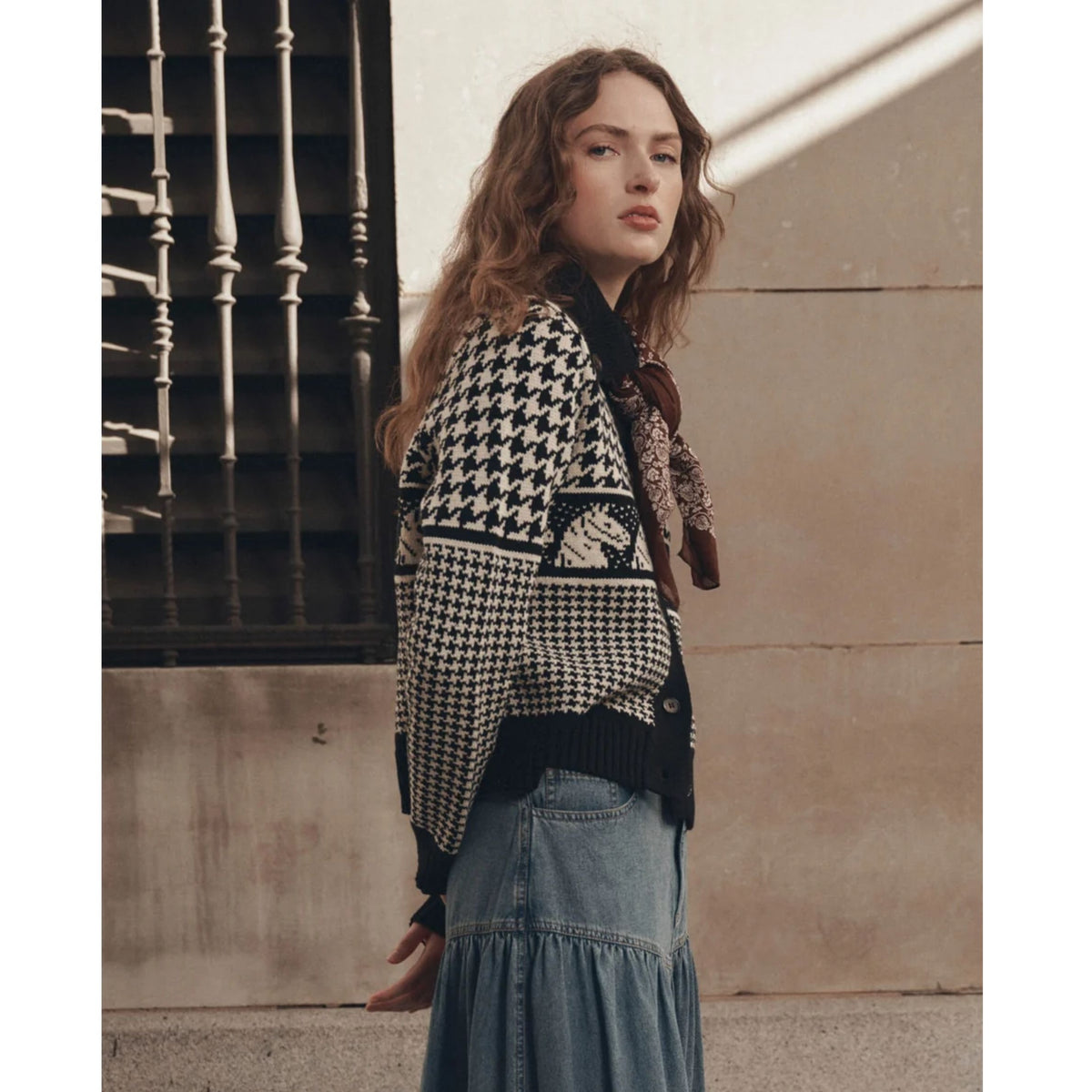 A woman wearing a black and white houndstooth patterned cardigan with a horse motif, paired with a denim maxi skirt, standing against a textured beige wall.