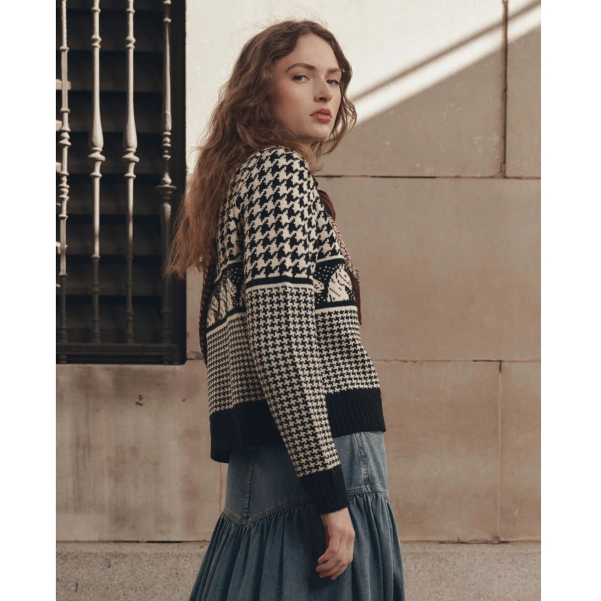 A woman wearing a black and cream houndstooth knit cardigan with a patterned yoke, paired with a blue tiered skirt, standing against a beige stone wall.