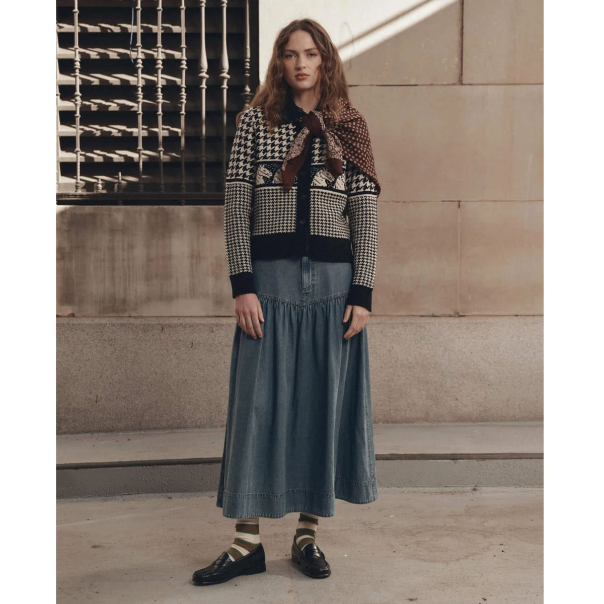 A model stands outdoors against a stone wall wearing a patterned knit cardigan, a long denim skirt, striped socks, and black loafers.
