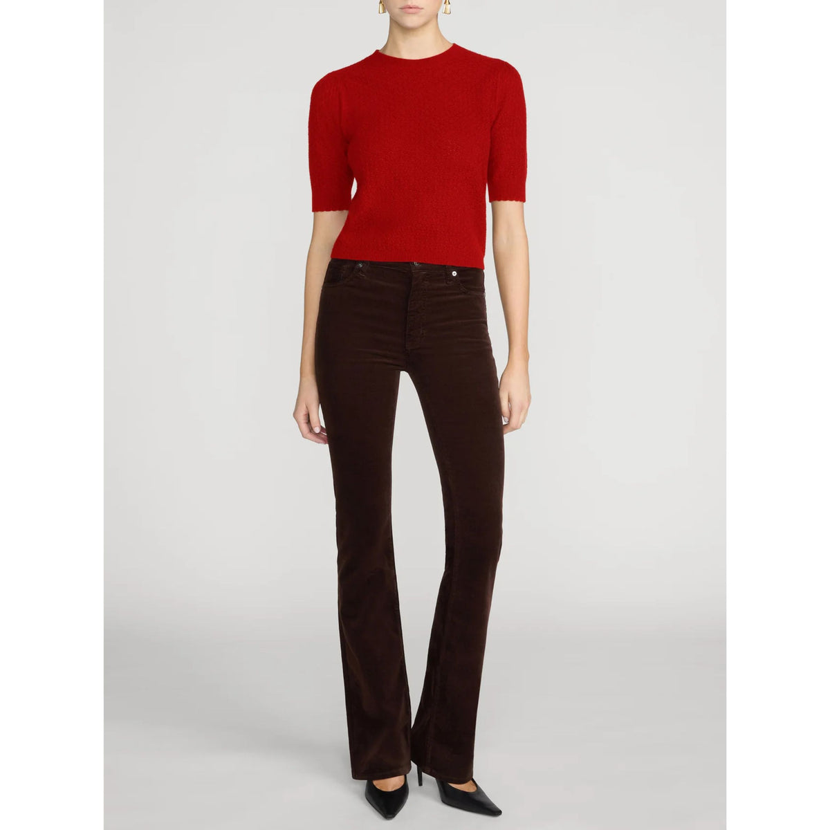 A model wearing a vibrant red short-sleeve knit top and dark brown flared velvet pants, paired with black pointed-toe heels, against a light gray studio background.