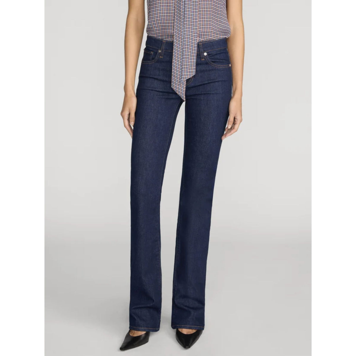 A model wearing dark blue straight-leg denim jeans, paired with a patterned shirt and black pointed shoes, shown from the waist down against a light gray studio background.