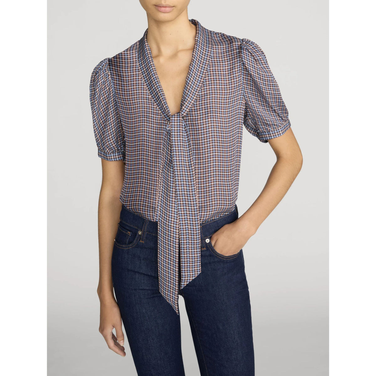 A woman wearing a short-sleeve plaid blouse with a tie-neck detail and dark blue jeans, photographed against a light gray studio background.