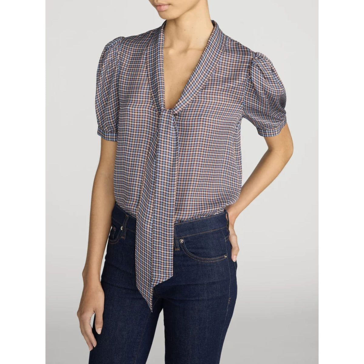 A woman wearing a short-sleeve plaid blouse with puff sleeves and a front tie detail, paired with dark blue jeans, against a light gray studio background.