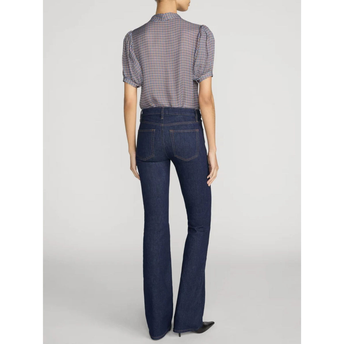 Rear view of a model wearing dark blue flared jeans and a short-sleeve checkered blouse against a light gray studio background.