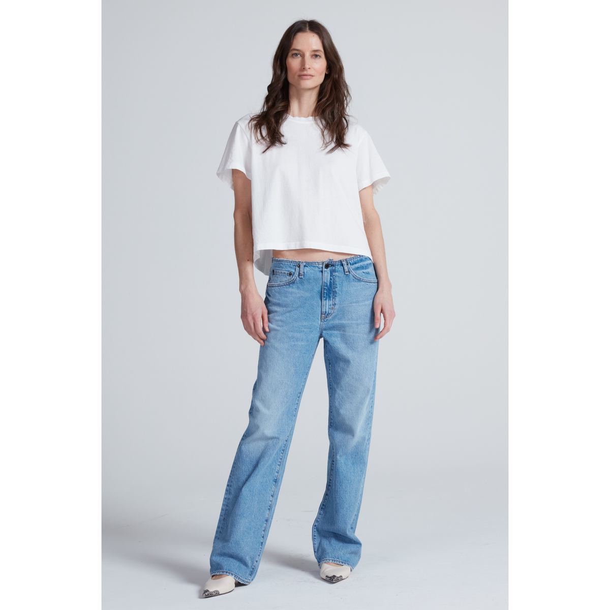 A woman modeling a white short-sleeve cropped T-shirt and light-wash blue wide-leg jeans against a light gray studio background.