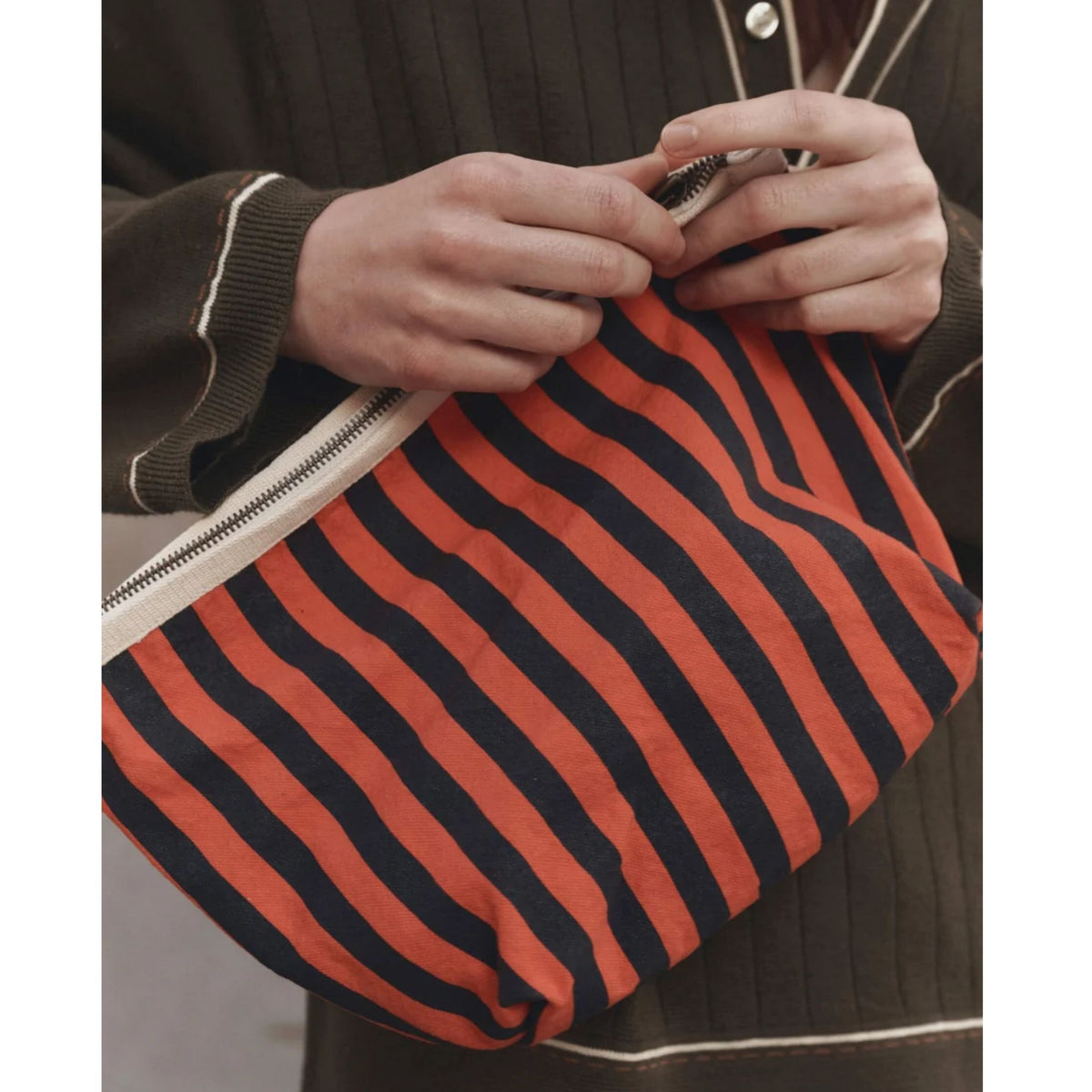 A close-up of a person holding a soft zip pouch with bold orange and black horizontal stripes. The person is wearing a dark brown ribbed cardigan with white piping.