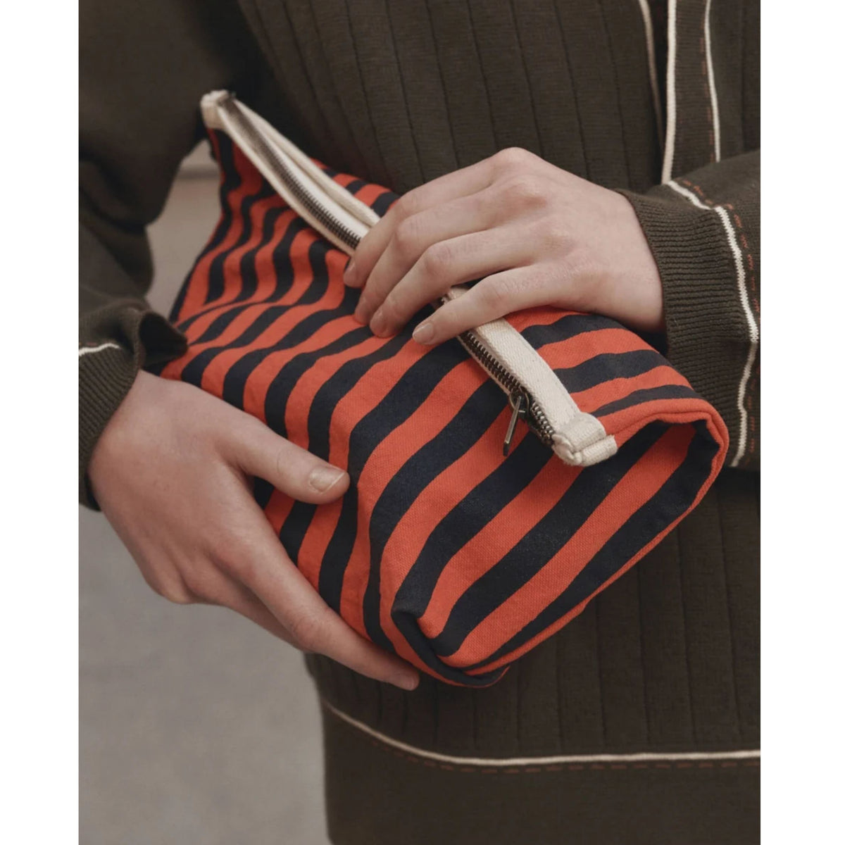 A close-up of a person holding a striped clutch bag with orange and black stripes and a cream zipper, wearing a dark ribbed sweater with white piping.