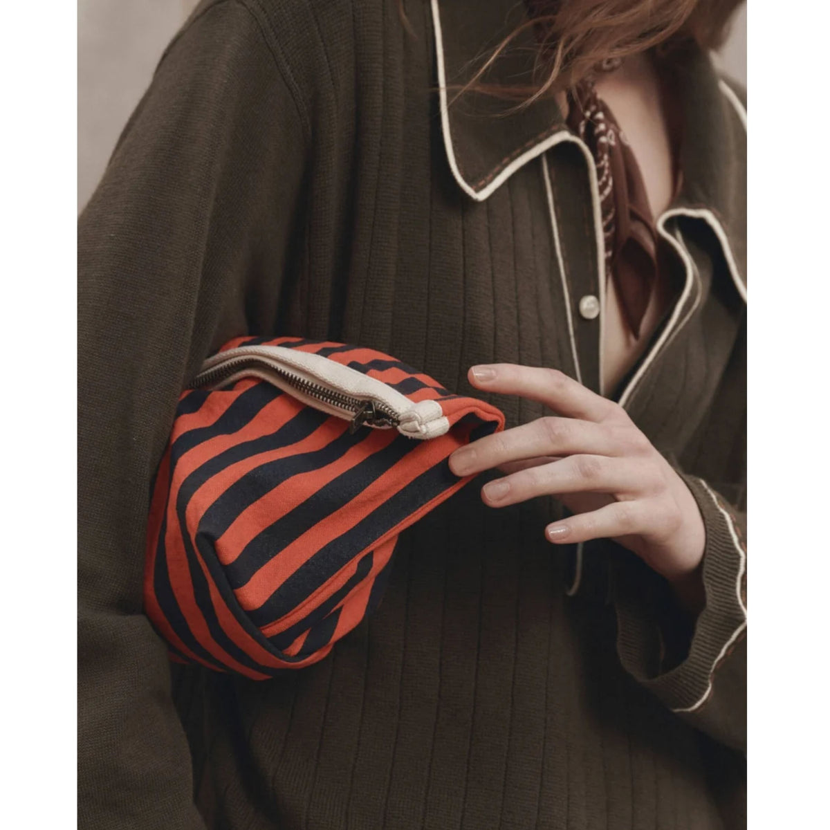 A close-up of a person wearing a dark olive ribbed jacket with white piping, holding a small zip pouch with bold orange and black stripes.