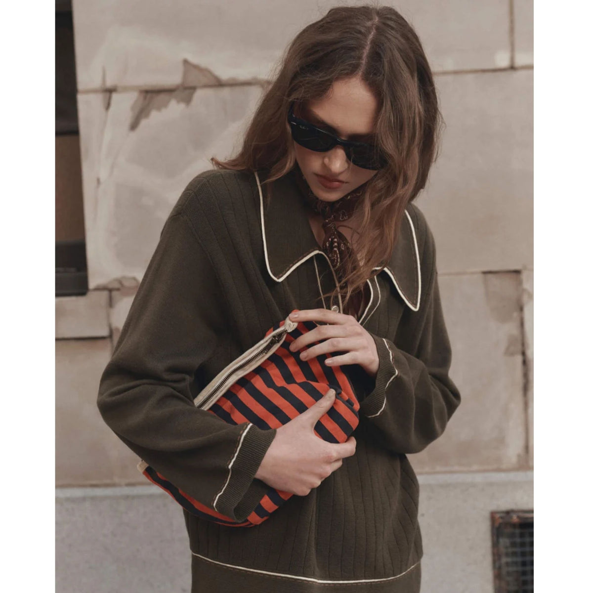 A woman wearing a dark olive green ribbed knit sweater with white piping and black sunglasses, holding a red and black striped clutch bag.