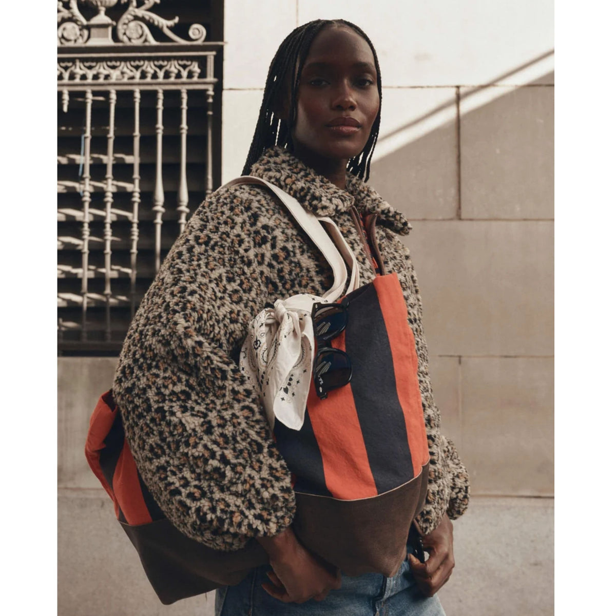 A woman wearing a leopard-print fuzzy jacket and blue jeans carries a large tote bag with orange, brown, and navy panels, a white scarf, and sunglasses. She is standing outdoors against a beige stone wall and black metal gate.