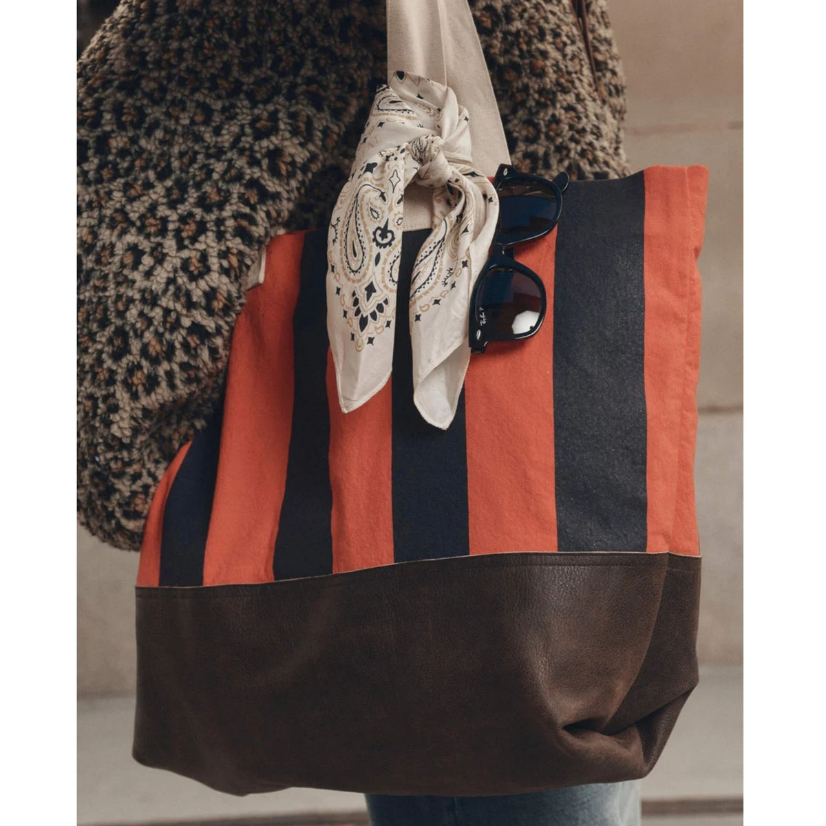 A striped tote bag with vertical orange, dark navy, and brown stripes, featuring a solid dark brown faux-leather bottom panel. A cream paisley bandana and black sunglasses are styled on top of the bag, and a leopard-print jacket is visible on the left side.