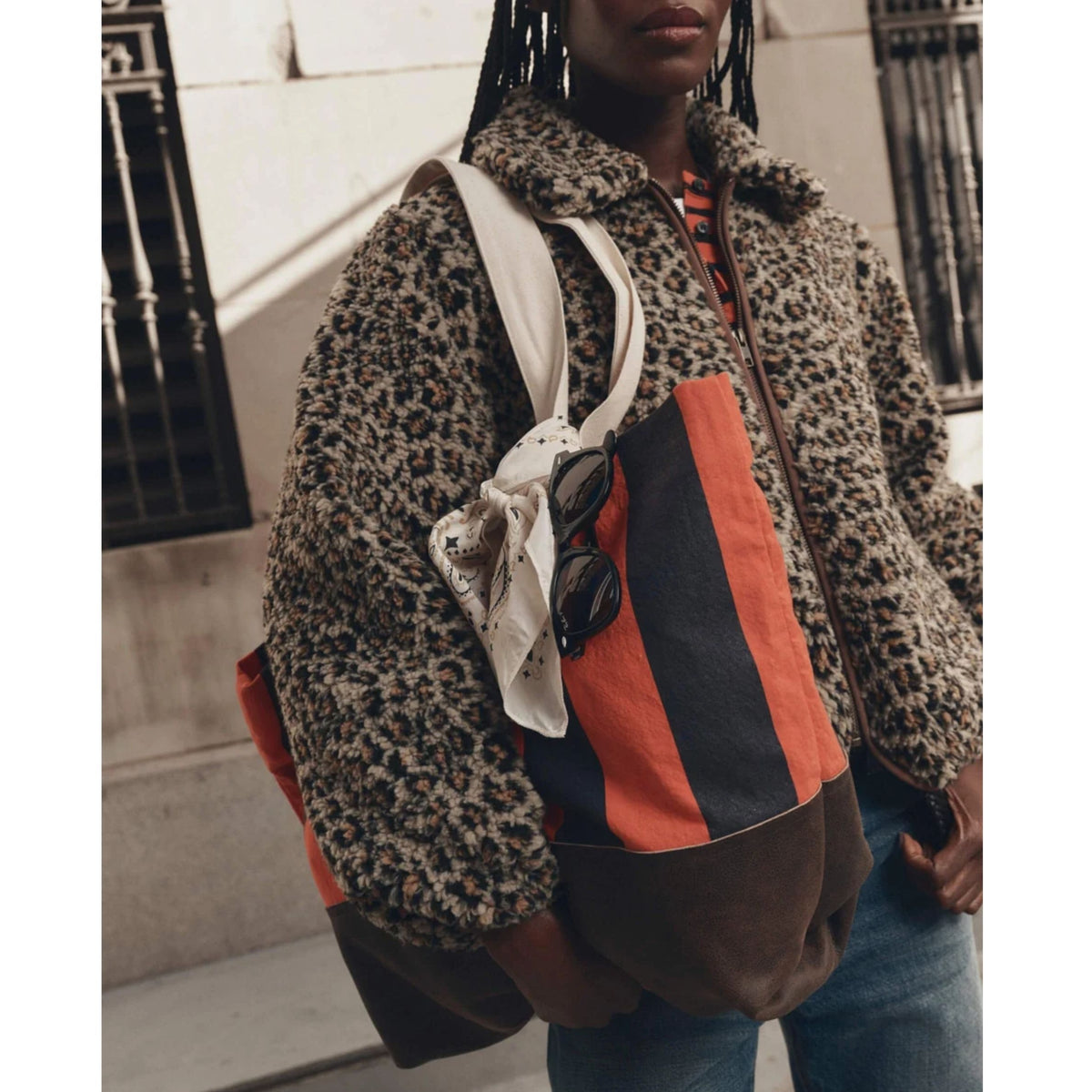 A person wearing a leopard-print fuzzy jacket and blue jeans carries a large shoulder bag with a brown base, orange and navy vertical stripes, and a white strap. Sunglasses and a white scarf are tucked into the strap.