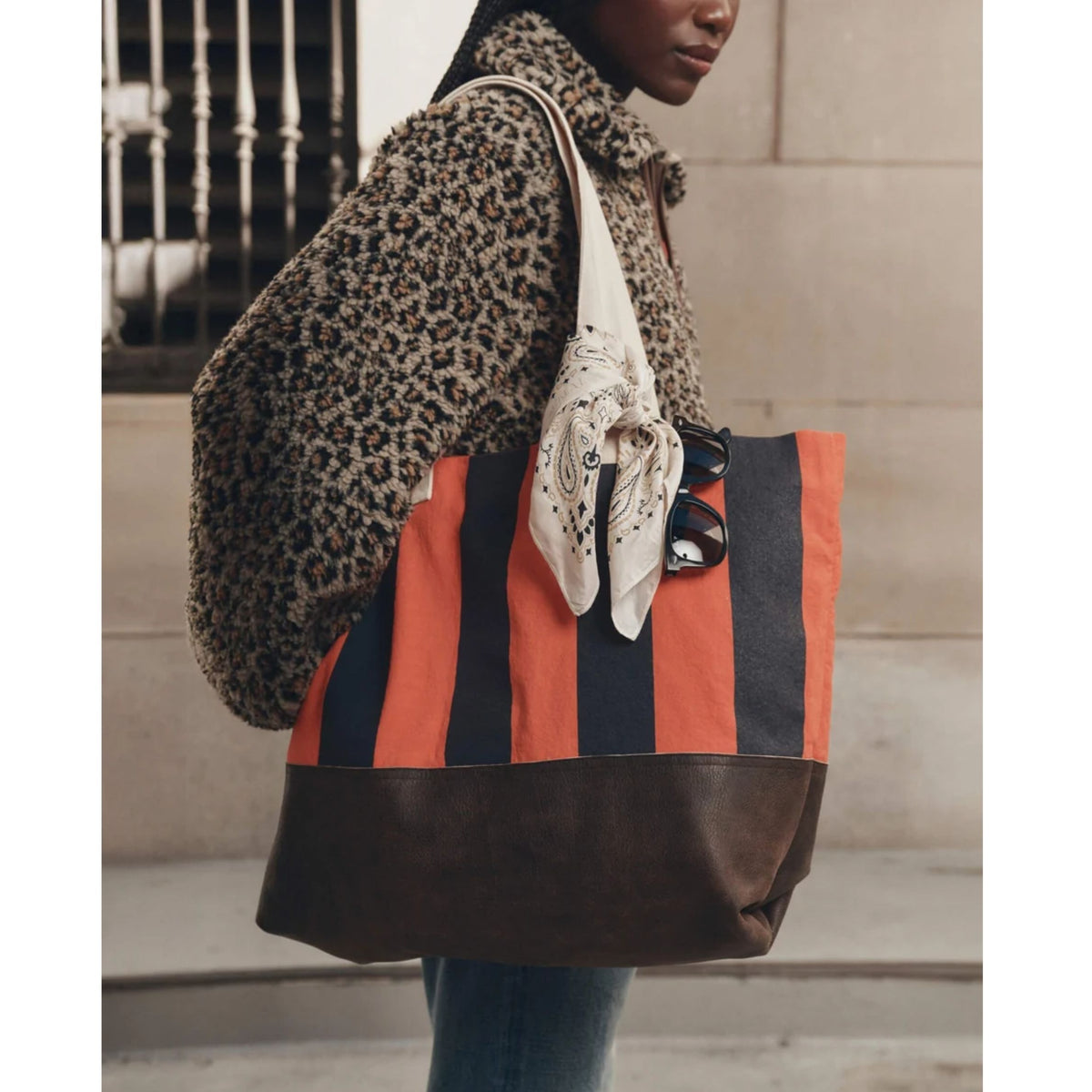 A person wearing a leopard-print coat carries a large striped tote bag with orange, navy, and dark brown vertical stripes and a dark brown leather base, styled with a cream scarf and sunglasses.