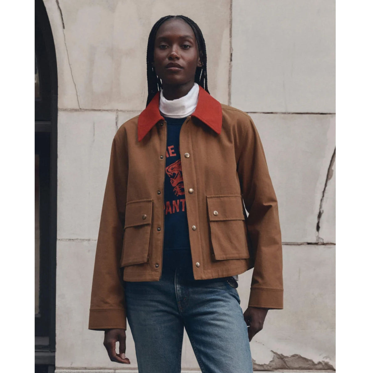 A model wearing a brown cropped utility jacket with a vibrant red collar, layered over a navy graphic t-shirt and blue jeans, standing against a textured light gray stone wall.