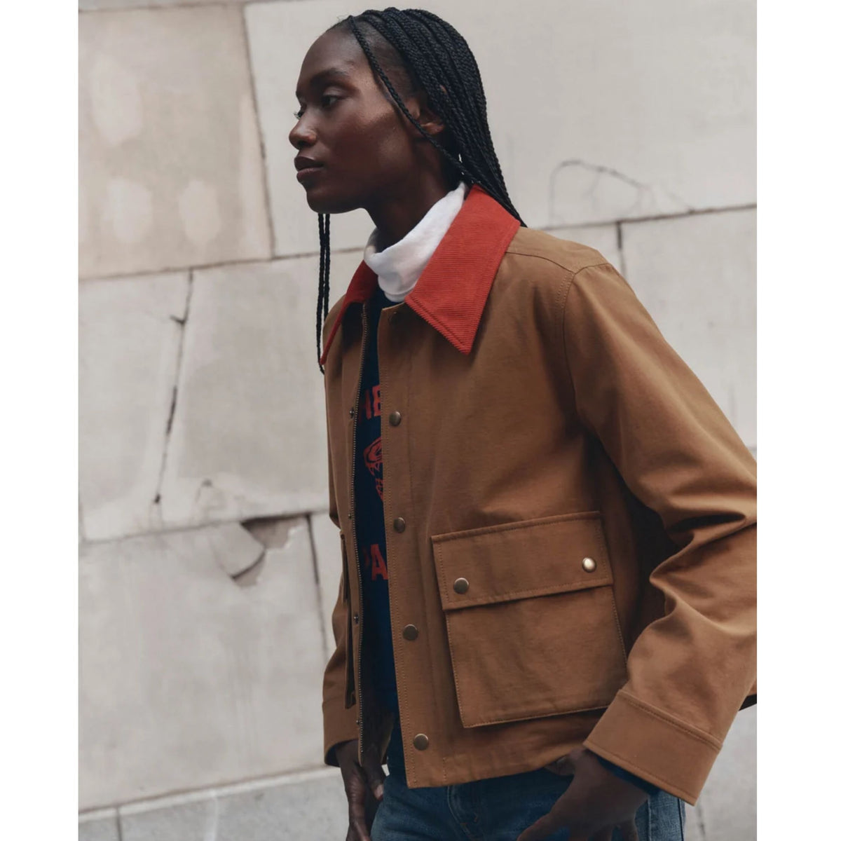 A woman wearing a brown utility jacket with a contrasting red-orange collar, layered over a white turtleneck and a dark graphic tee, set against a textured concrete wall.