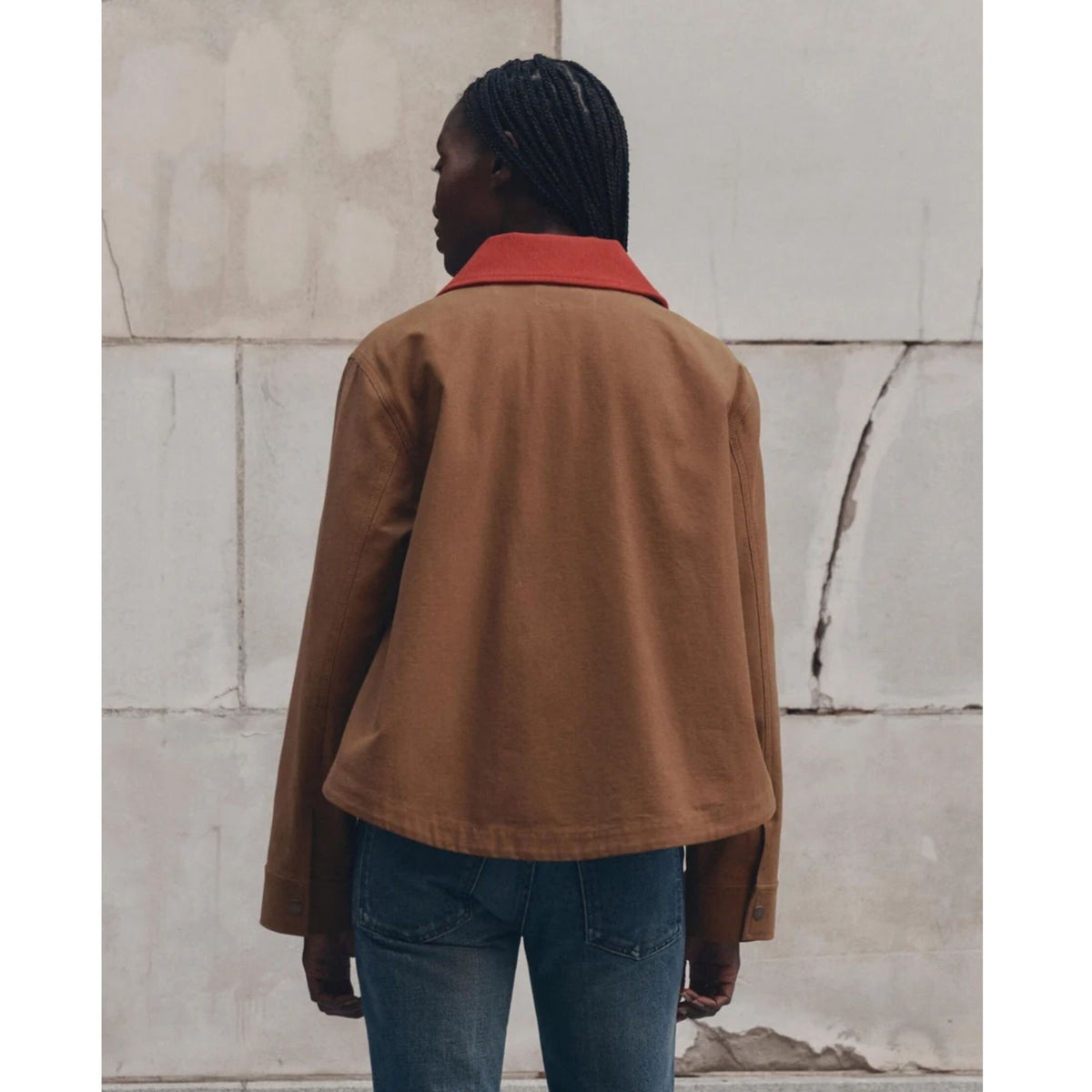 A model viewed from behind wearing a cropped brown jacket with a contrasting red collar, paired with blue jeans, standing against a light gray stone wall.