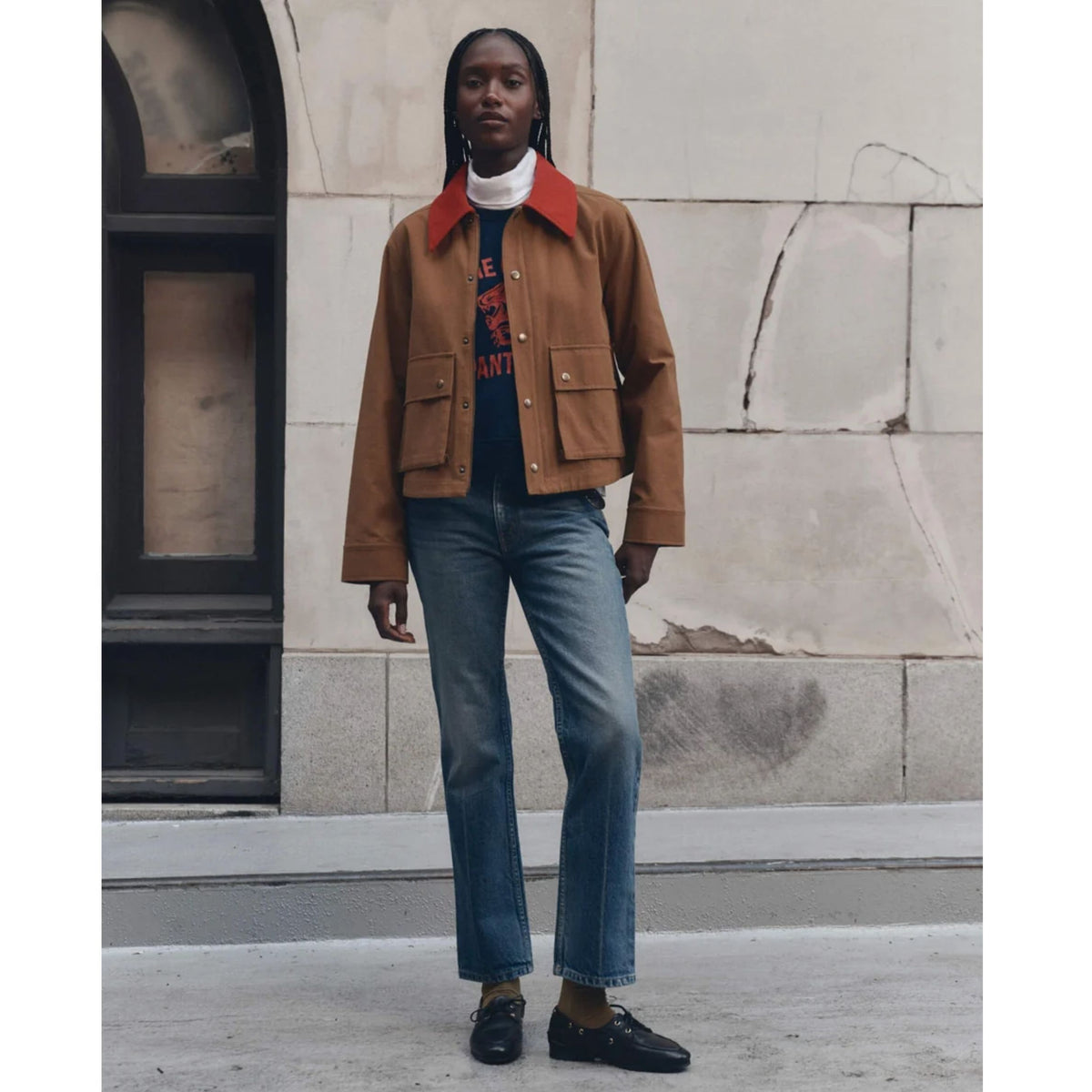 A model stands on a city sidewalk wearing a brown cropped jacket with a contrasting red collar, blue jeans, a black graphic t-shirt, and black loafers, set against a stone building background.