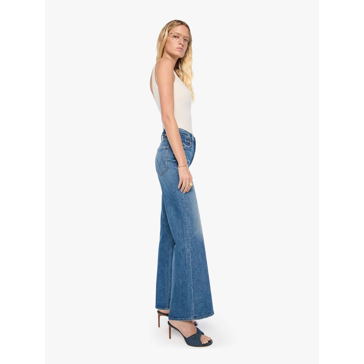 A woman modeling a pair of high-waisted, wide-leg blue denim jeans with a white sleeveless top and dark blue heeled sandals, shown from a side profile against a light gray studio background.