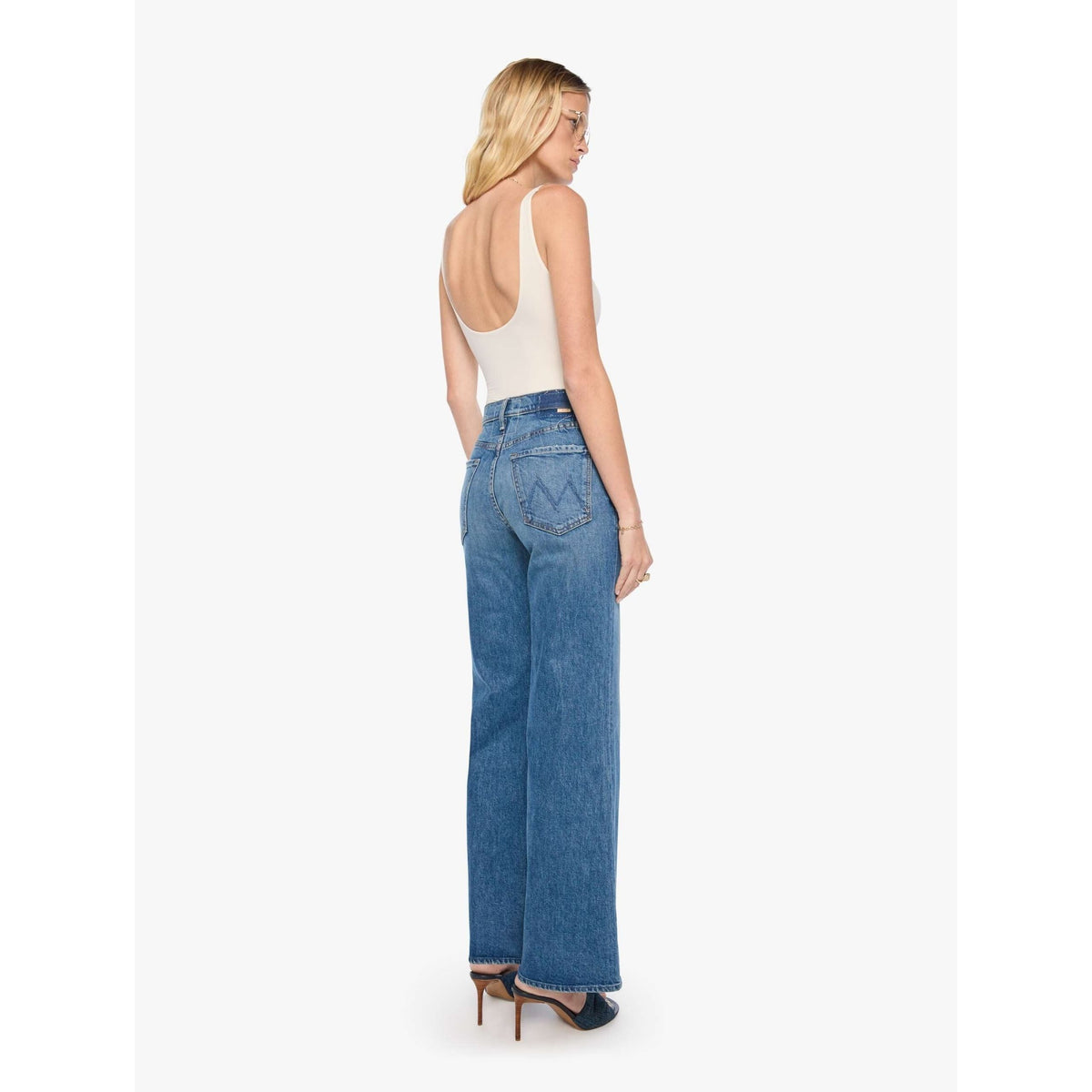 A woman shown from the back wearing blue wide-leg jeans, a white sleeveless top, and dark blue heels against a plain light gray studio background.