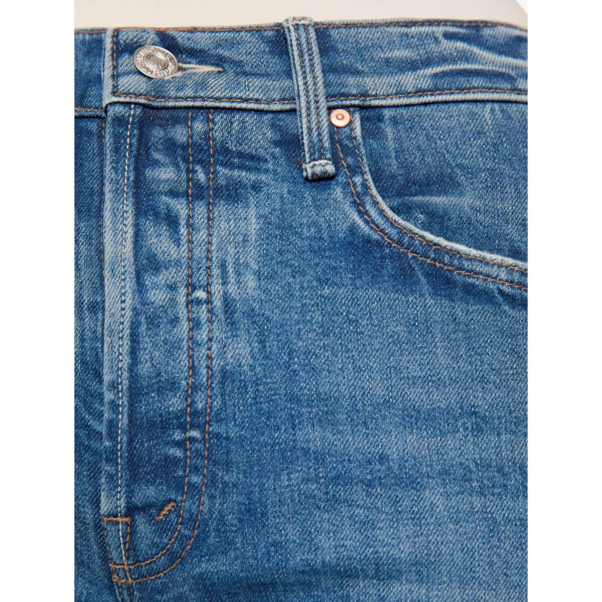 Close-up of blue denim jeans showing the waistband, belt loops, button, rivet, front pocket, and stitching.