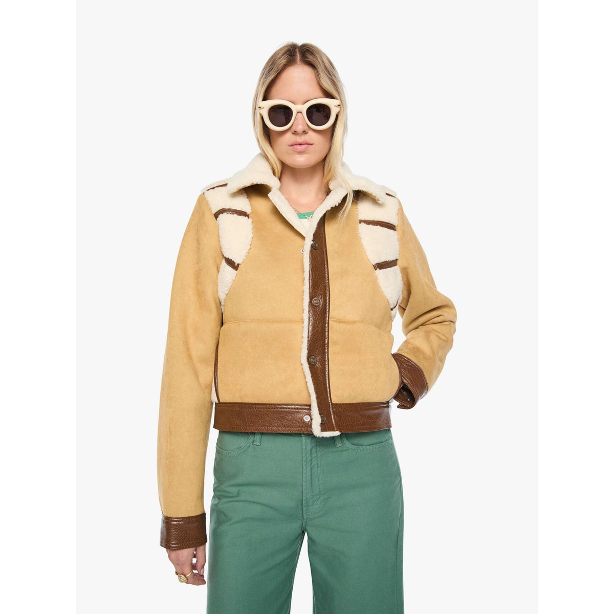A woman modeling a cropped tan shearling jacket with brown faux-leather trim and a cream lining, paired with green pants and round sunglasses, against a plain white background.