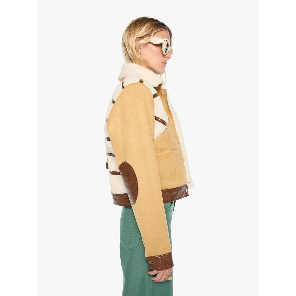 A woman shown in side profile wearing a cropped shearling-style jacket with tan suede-like panels, cream fleece lining, and brown trim, paired with green pants against a plain light gray studio background.