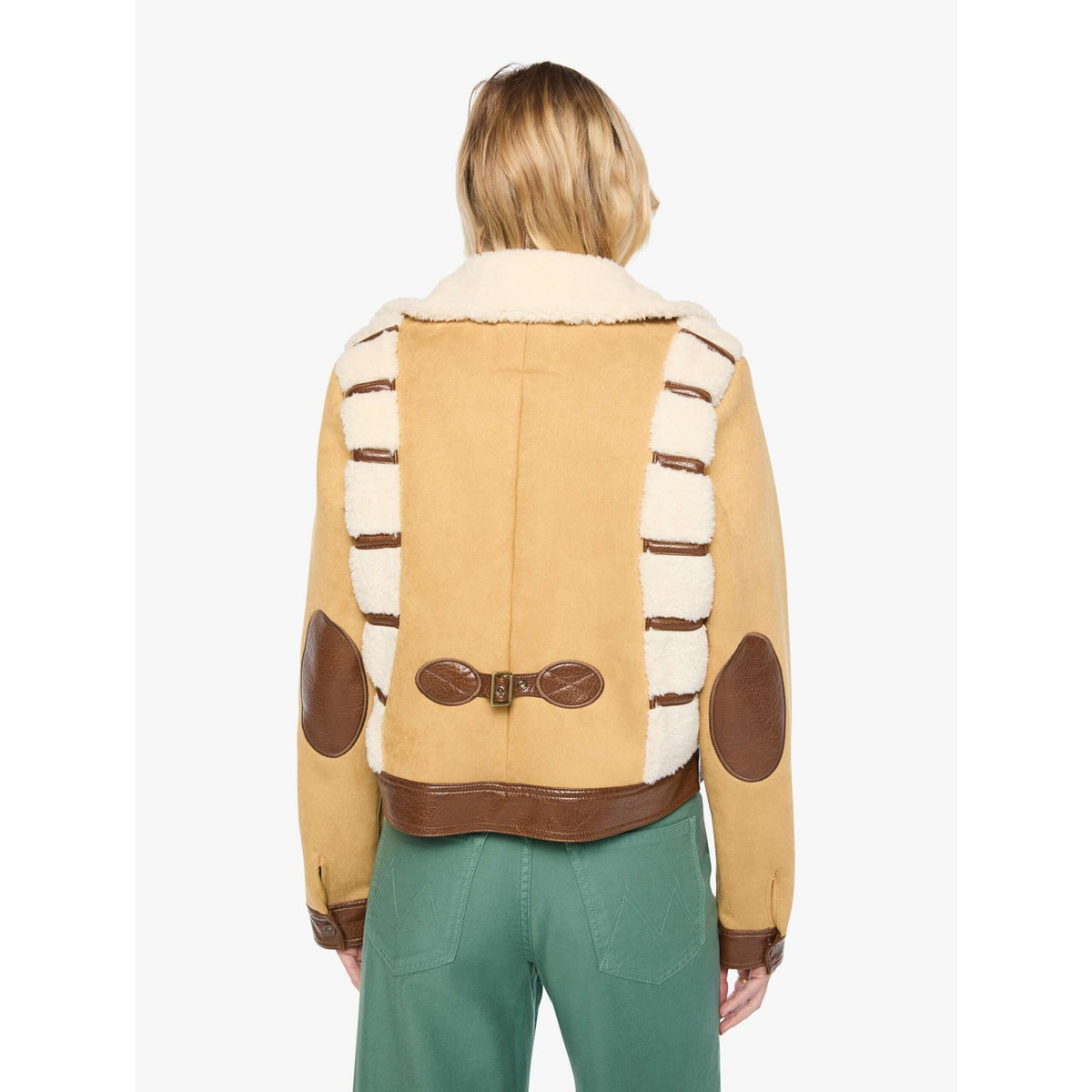 A model viewed from the back wearing a tan shearling jacket with brown leather trim and white-and-brown striped shoulder panels, paired with green pants, against a plain light gray studio background.