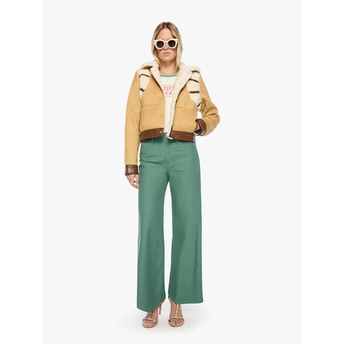 A model wearing a tan shearling-lined aviator jacket with brown leather trim, paired with wide-leg green trousers, a graphic t-shirt, and sunglasses, standing against a plain white studio background.