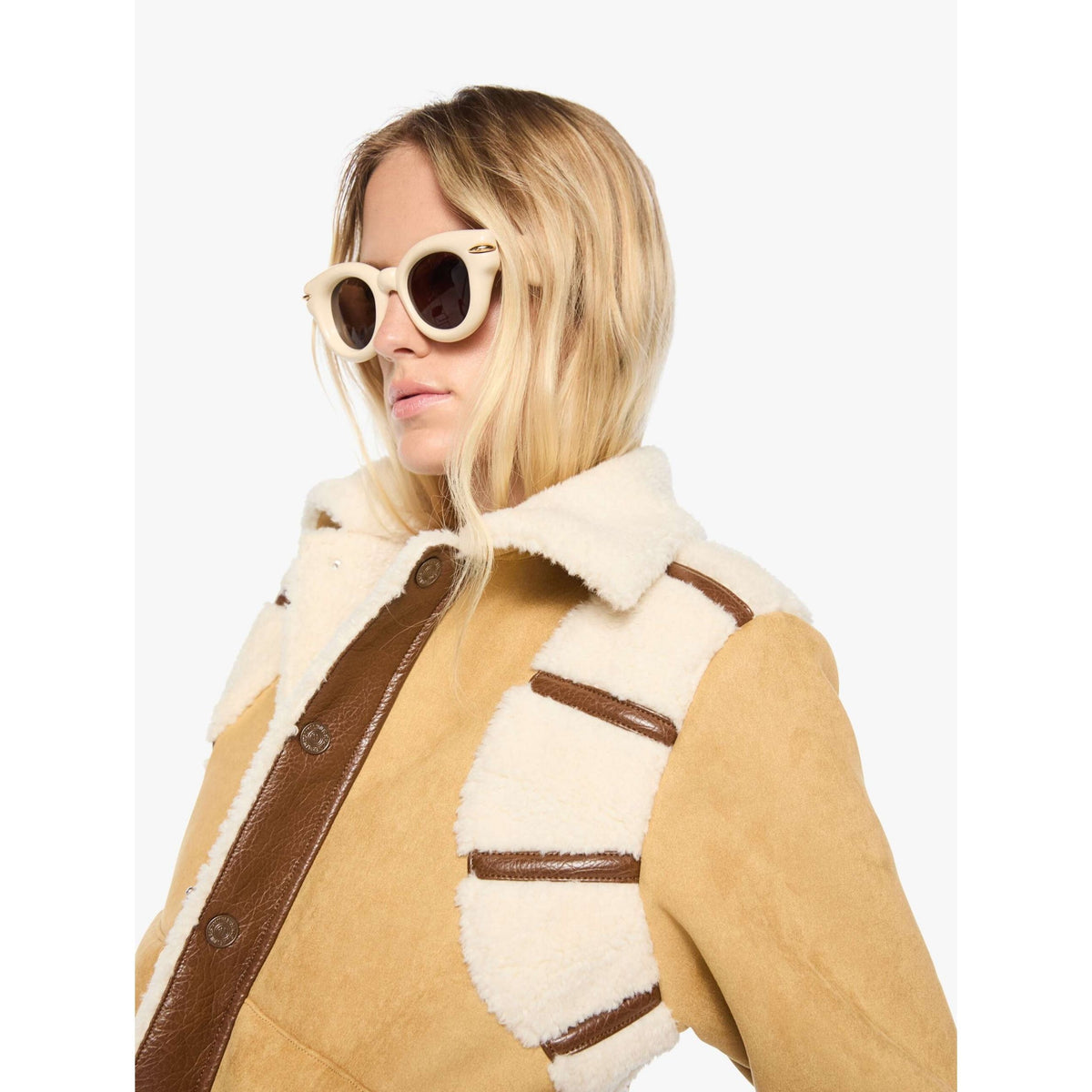 A woman wearing a tan and cream shearling-style jacket with brown trim, photographed against a plain light gray studio background. She is also wearing cream-framed sunglasses.
