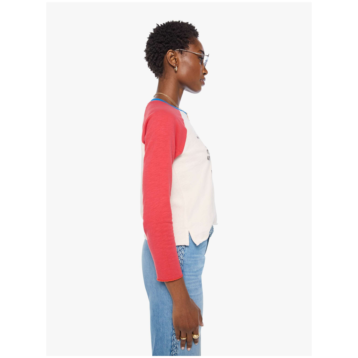 A side-profile view of a model wearing a color-blocked knit top with a coral red three-quarter sleeve and body, and an off-white front panel, paired with light blue jeans against a plain light gray background.