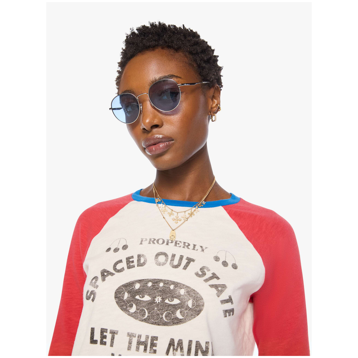A model wearing round sunglasses and a red and white raglan graphic t-shirt with blue collar trim, set against a plain light gray background.