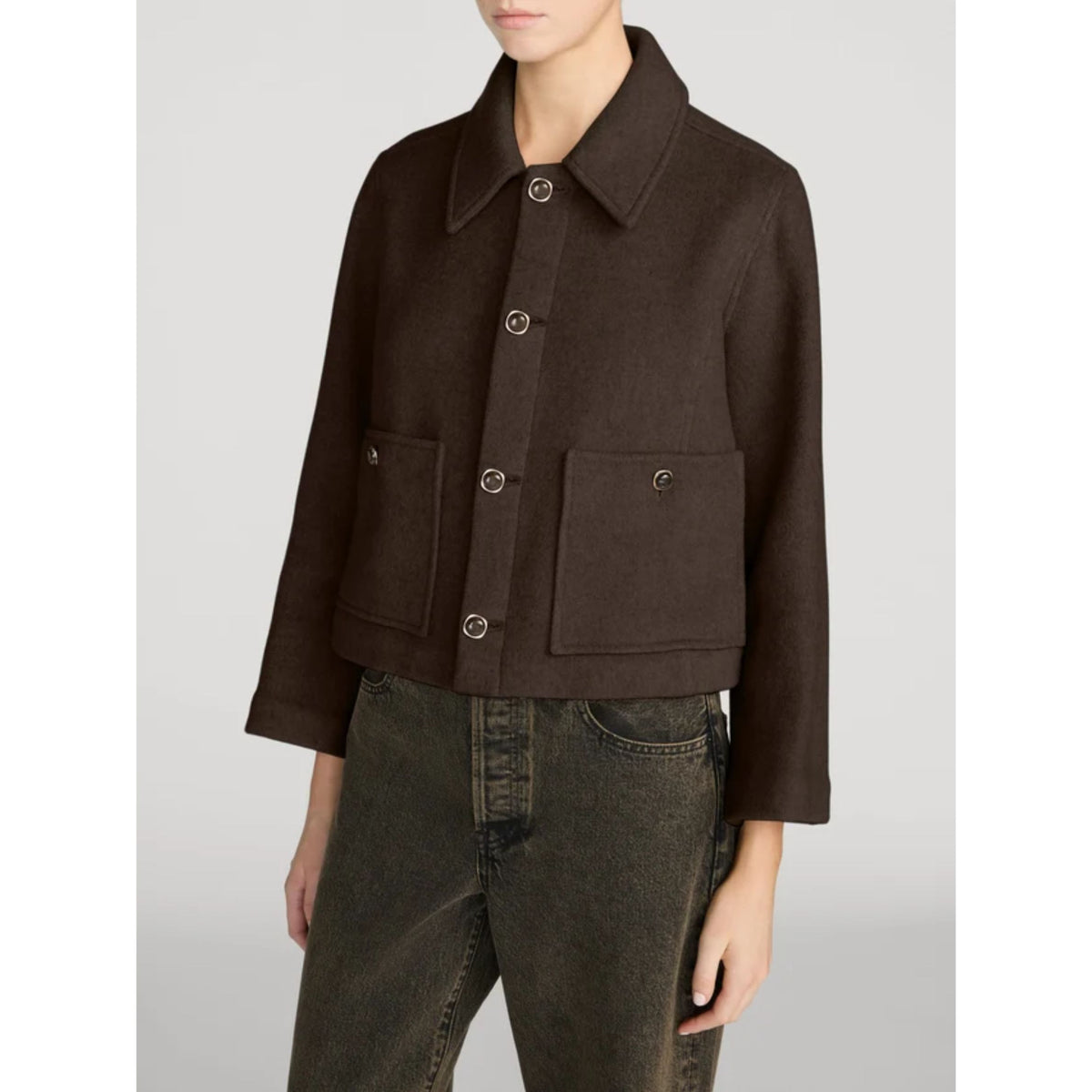 A model wearing a dark brown cropped jacket with a pointed collar, three front buttons, and two large front flap pockets, paired with dark gray jeans against a light gray studio background.