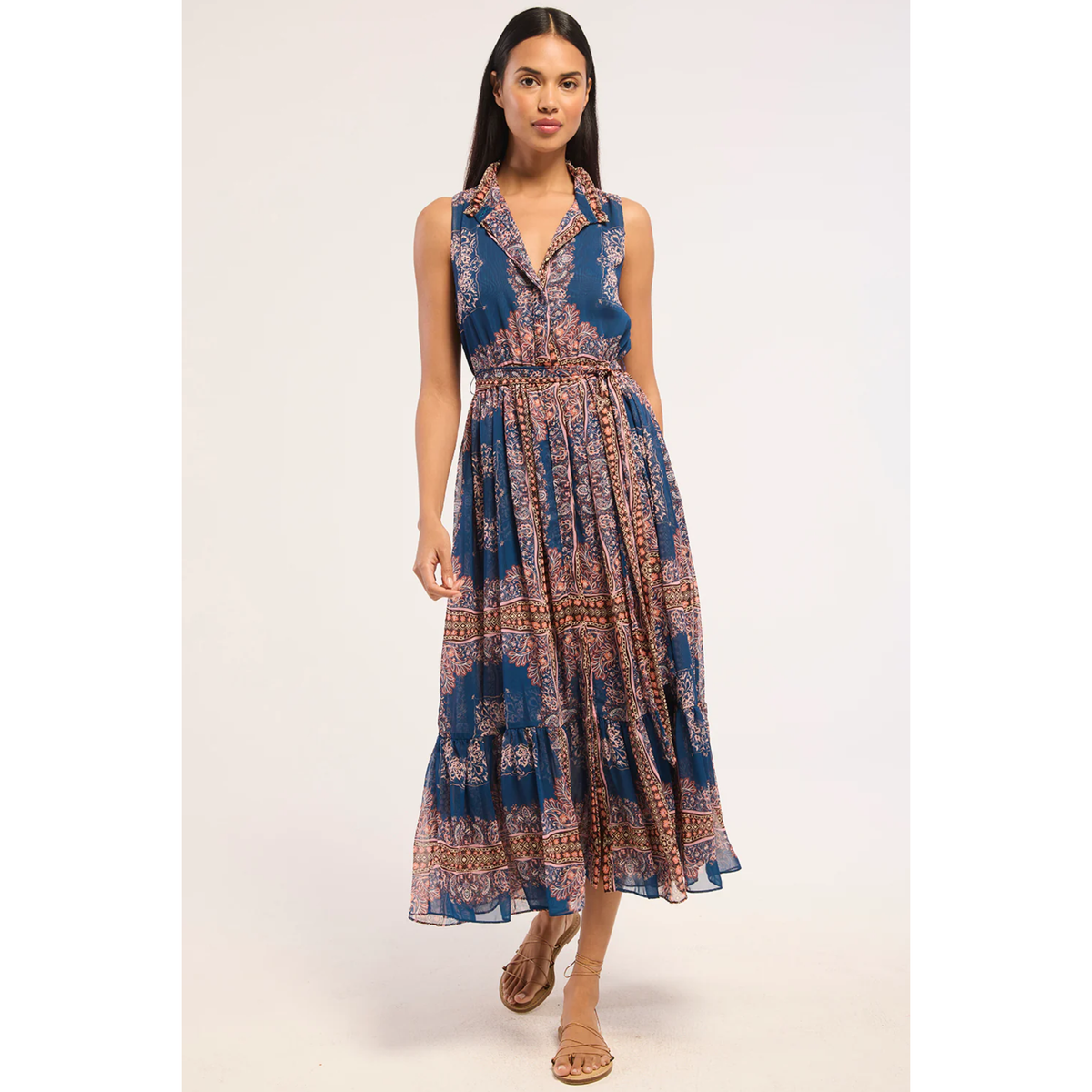A woman modeling a sleeveless midi dress with a blue base and a detailed brown and beige bohemian pattern, set against a plain light background.