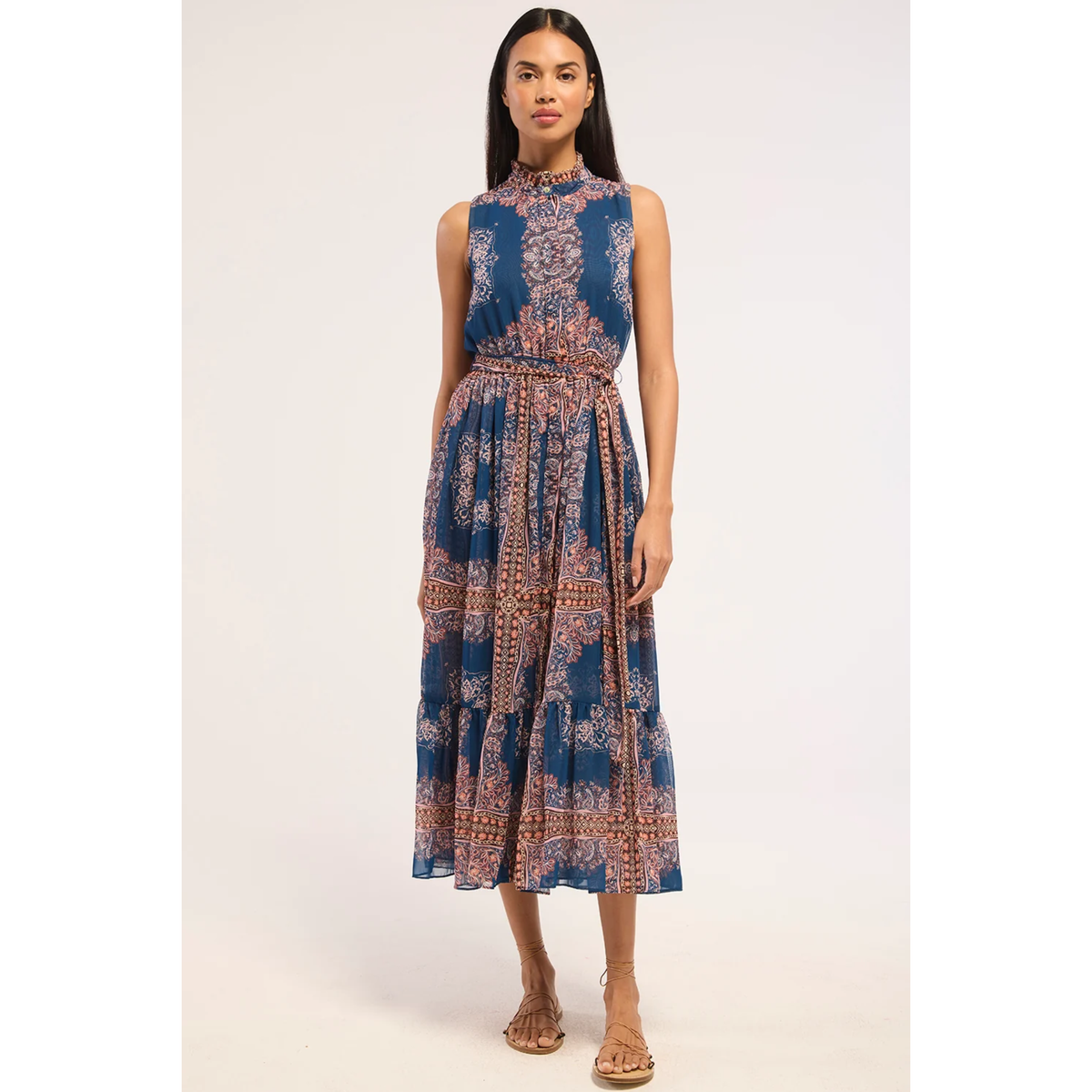 A woman modeling a sleeveless high-neck midi dress with a blue base and intricate rust-orange and beige paisley patterns, paired with tan strappy sandals against a plain light background.