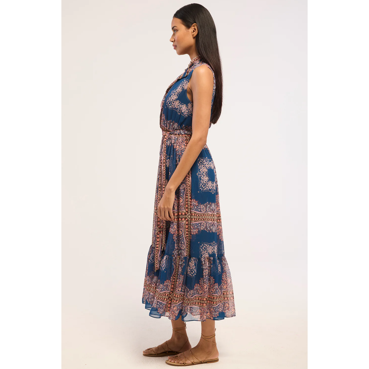 A woman modeling a sleeveless halter-neck midi dress with a blue base and intricate orange, beige, and white paisley patterns, shown in profile against a light gray studio background.