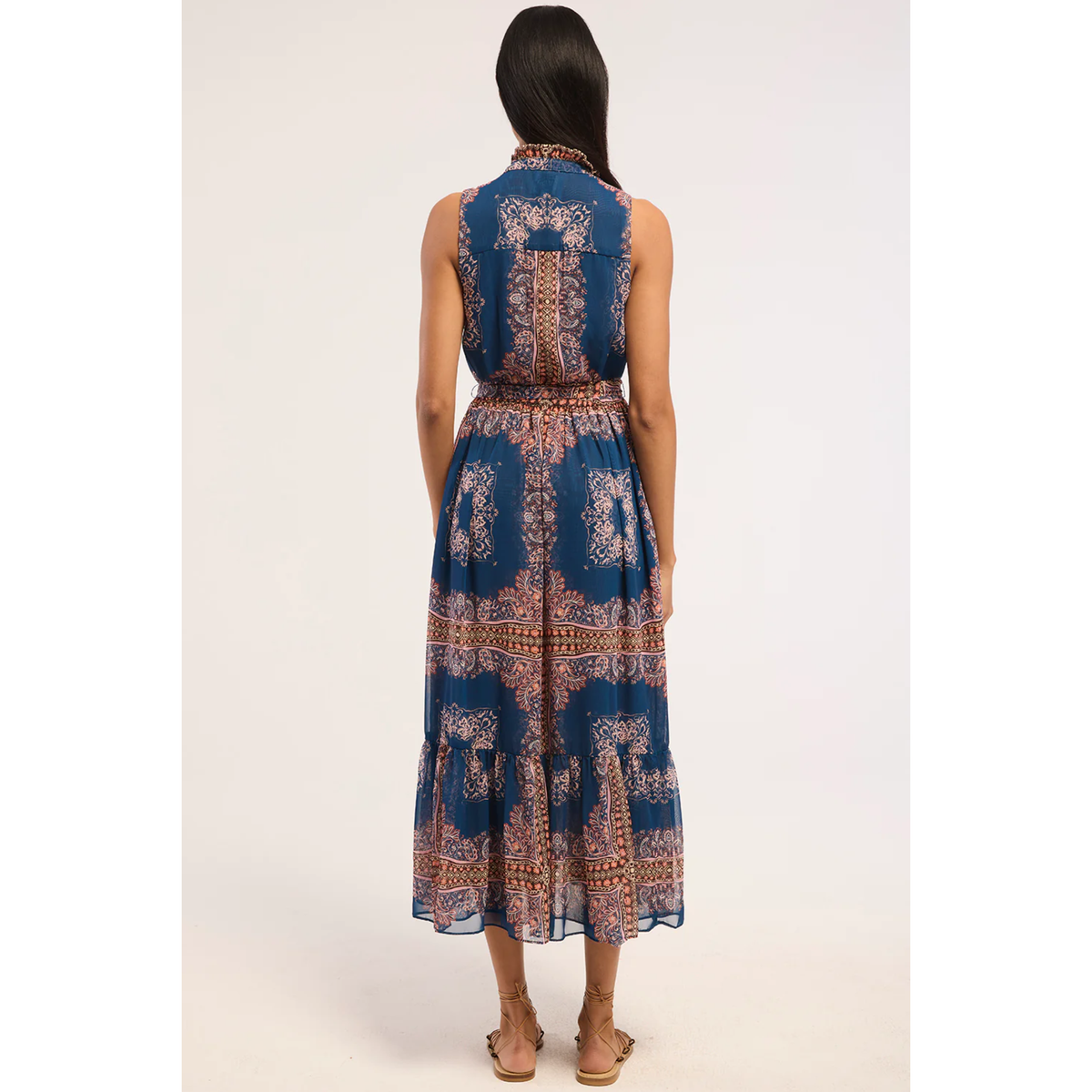 A woman viewed from the back wearing a sleeveless midi dress with a blue base and intricate brown and beige paisley-style print, paired with tan flat sandals against a plain light background.