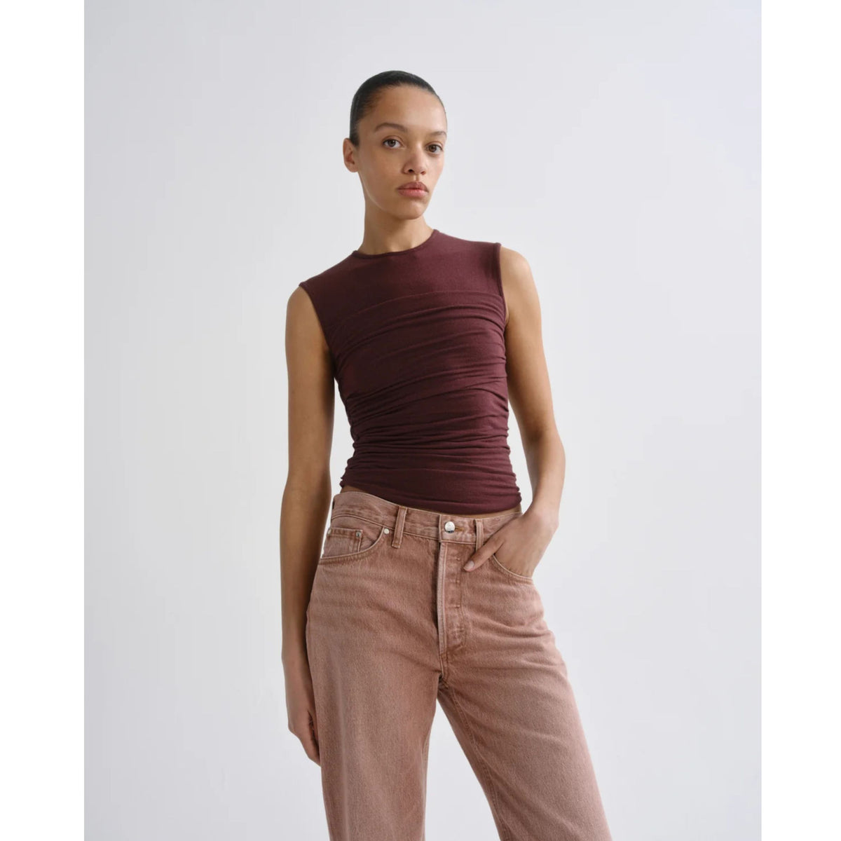 Model wearing the AGOLDE Theodore Tank in Currant—a sleeveless, fitted deep burgundy top with gathered waist detailing—paired with dusty pink pants against a light gray background.