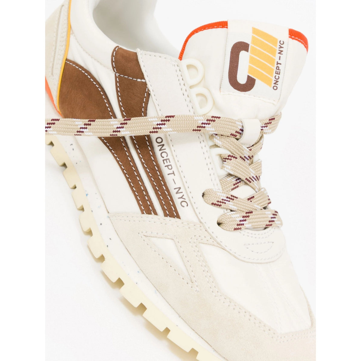 A close-up of a Oncept NYC sneaker in off-white suede and brown leather, featuring beige laces with red stripes and a yellow and brown logo on the tongue.