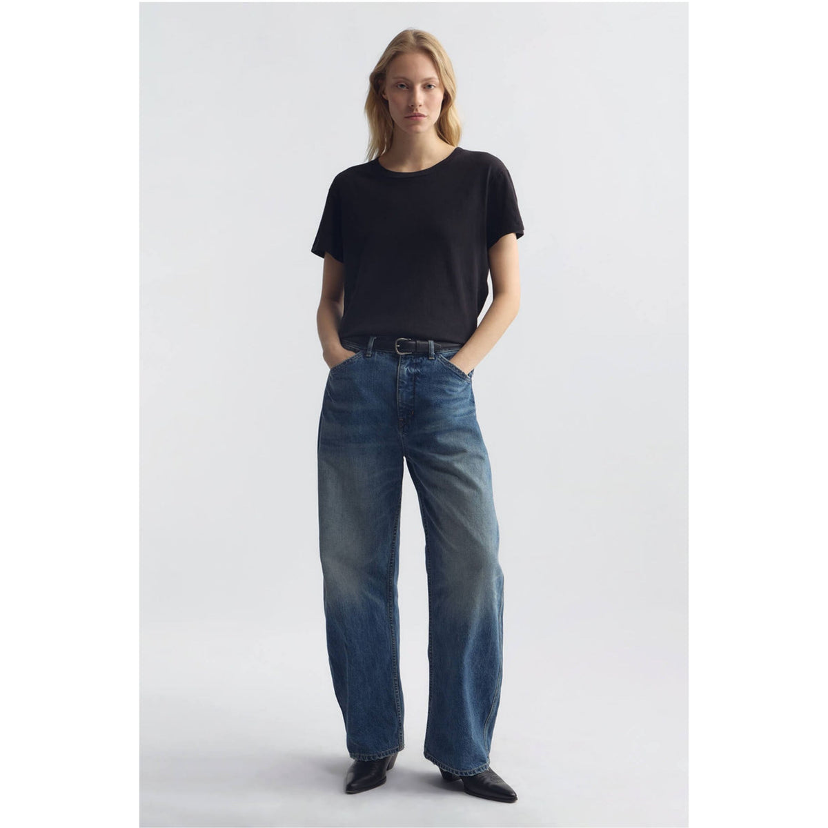 A woman stands against a light gray studio background wearing a black short-sleeve crew-neck t-shirt, wide-leg blue jeans, and black pointed shoes.